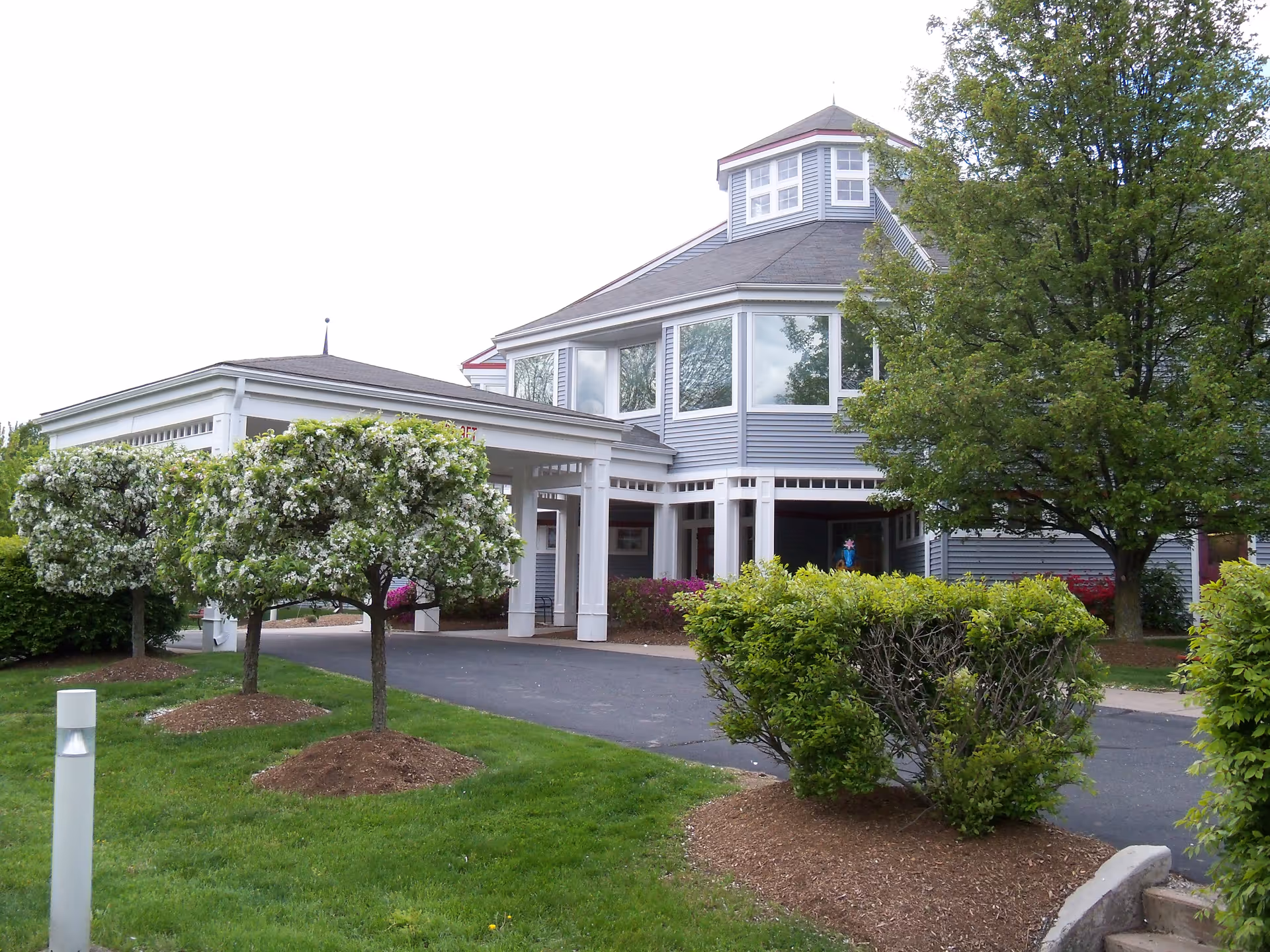 Exterior view of a senior living facility building with a covered entrance, surrounded by green grass, blooming trees, and shrubs.
