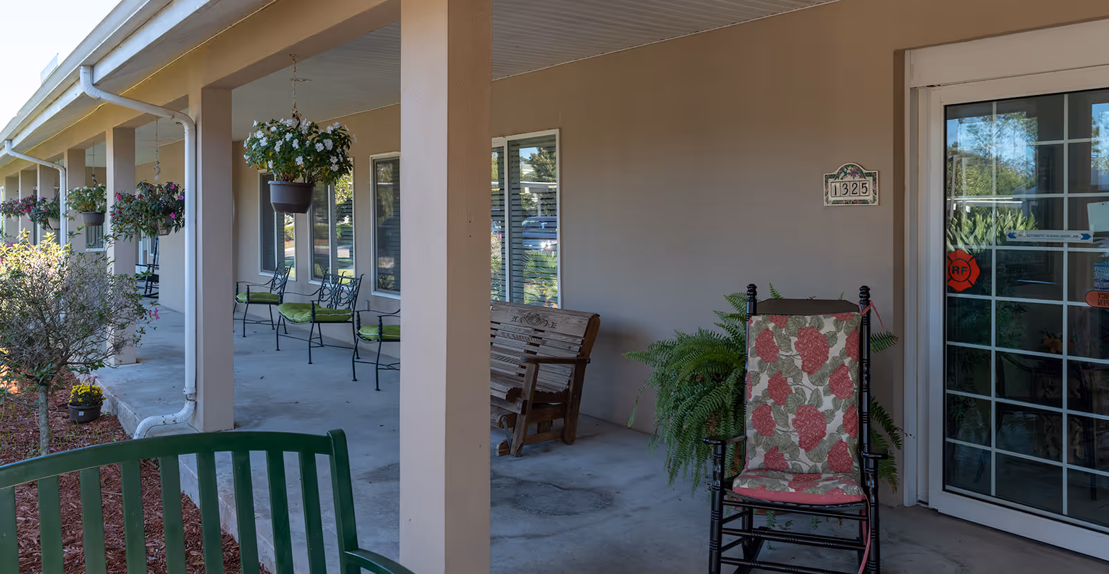 Covered outdoor patio area at Azalea Park senior living community with hanging flower pots, green cushioned metal chairs, a wooden bench, a rocking chair with floral cushions, and a sliding glass door entrance.