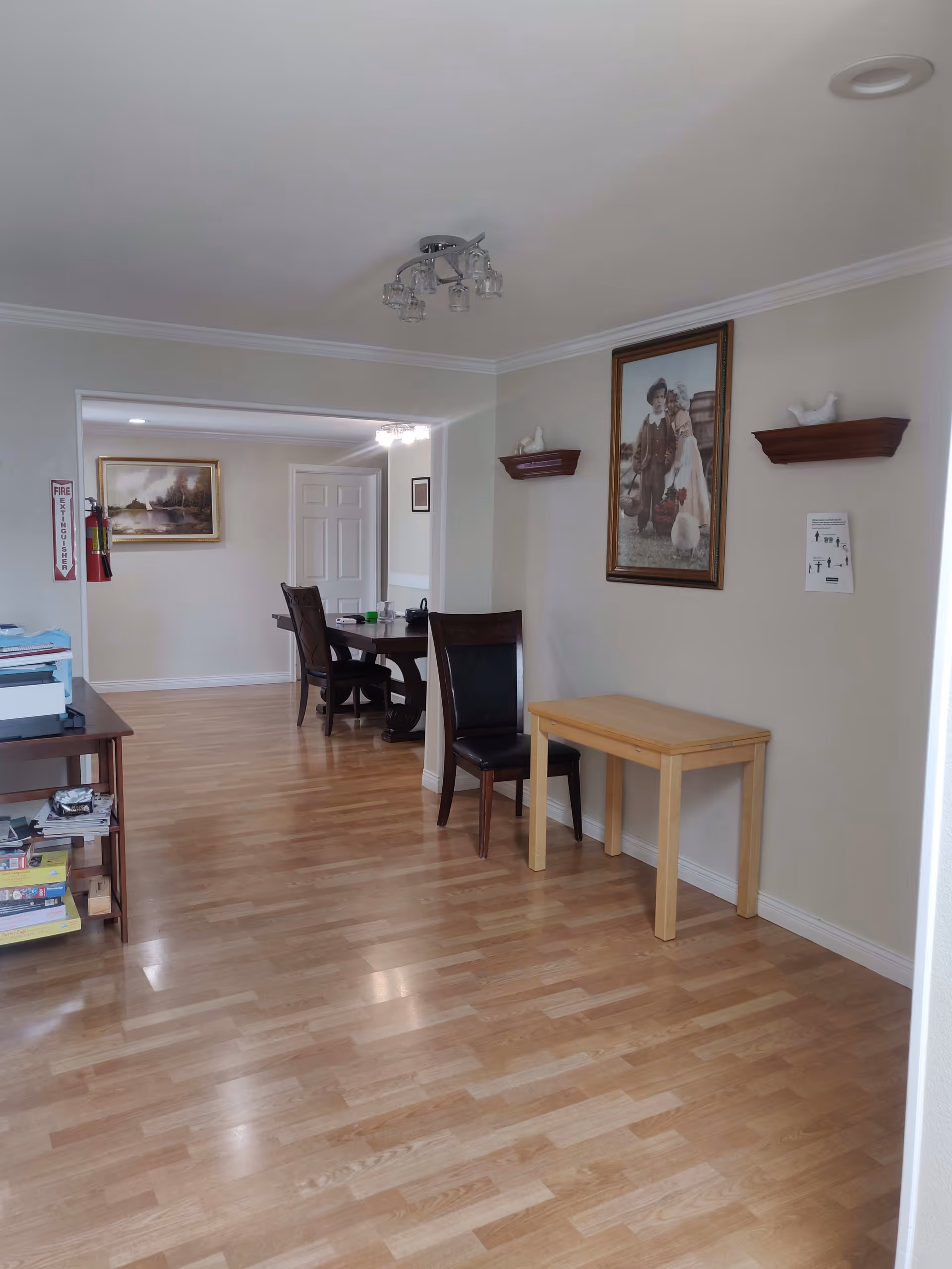 Interior view of a senior care facility showing a hallway with light wood flooring and beige walls. There is a small wooden table with a chair against the right wall, which has a framed picture of two children and two small shelves with decorative white bird figurines. Further down the hallway, there is a dining area with a dark wood table and chairs. A fire extinguisher and a framed landscape painting are visible on the left wall.