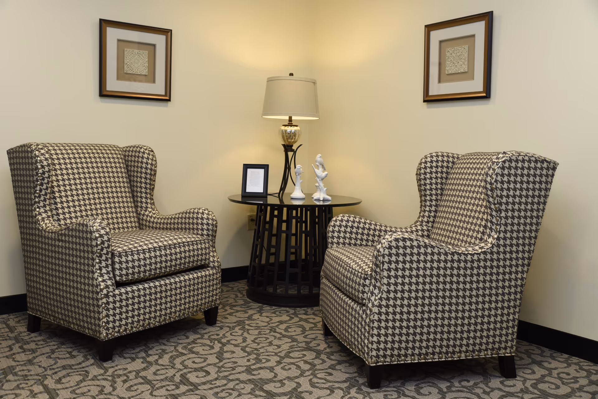 Two patterned wingback chairs facing each other with a round side table, lamp, and framed artwork on the wall in a small seating area.