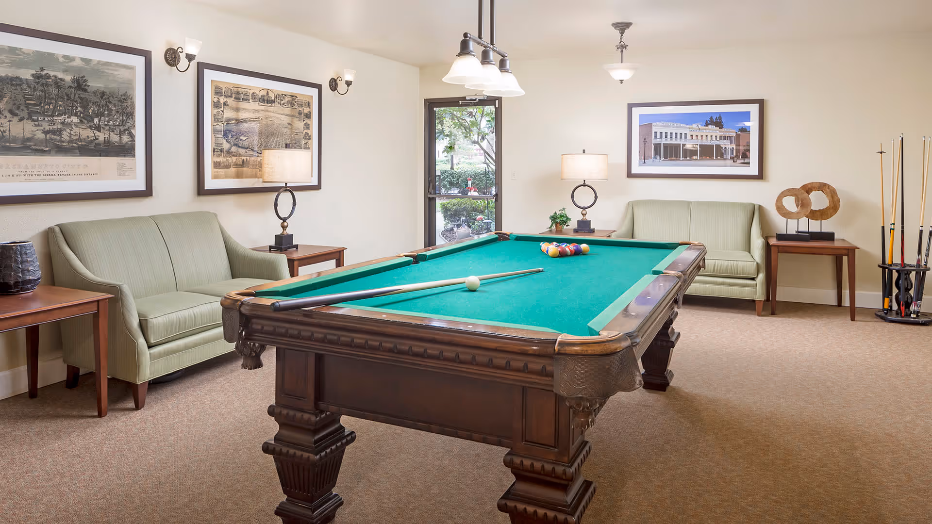 A well-lit room featuring a green felt pool table with cues and balls arranged for a game. The room has two light green sofas on either side of the pool table, each accompanied by a wooden side table with lamps. The walls are decorated with framed pictures, and there is a door with a window showing greenery outside. A rack with pool cues stands in the corner.