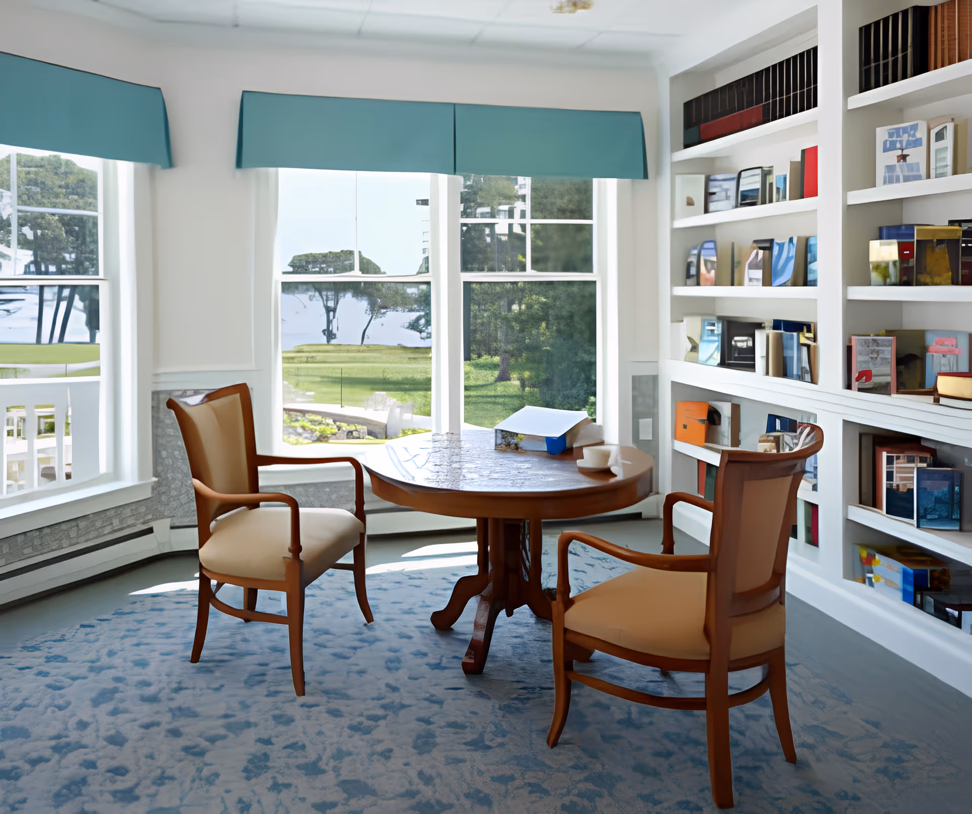 A bright room with large windows overlooking a green outdoor area. Inside, there is a round wooden table with two cushioned wooden chairs on a blue patterned carpet. A built-in white bookshelf filled with various books and decorative items lines one wall.