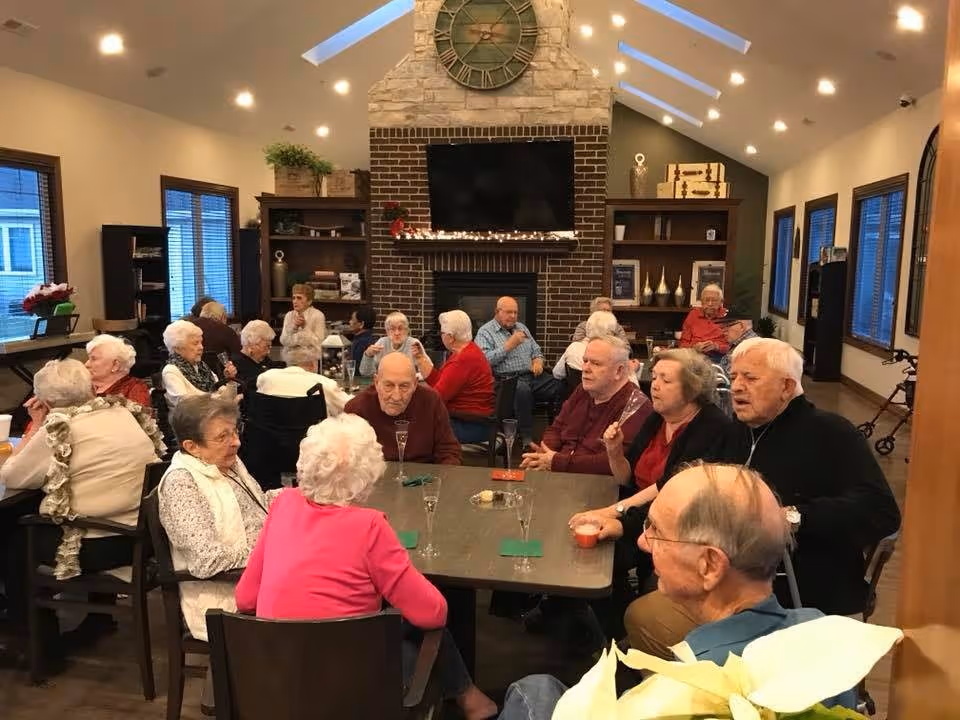 A group of elderly residents seated around tables in a cozy common room with a fireplace and television.