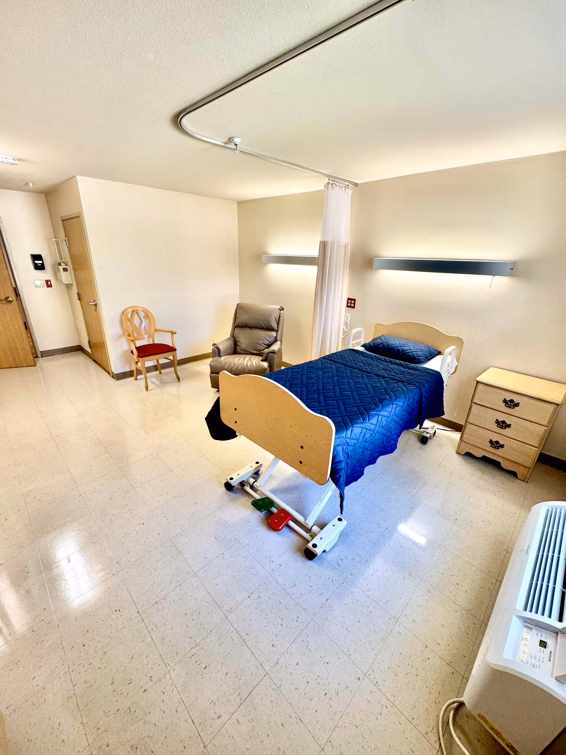 A simple, clean hospital or care facility room with a single bed covered in a blue quilt, a beige recliner chair, a wooden chair with a red cushion, a small wooden nightstand with three drawers, and a white air conditioning unit. The room has beige walls, a tiled floor, and a ceiling-mounted curtain track with a partially drawn white privacy curtain.