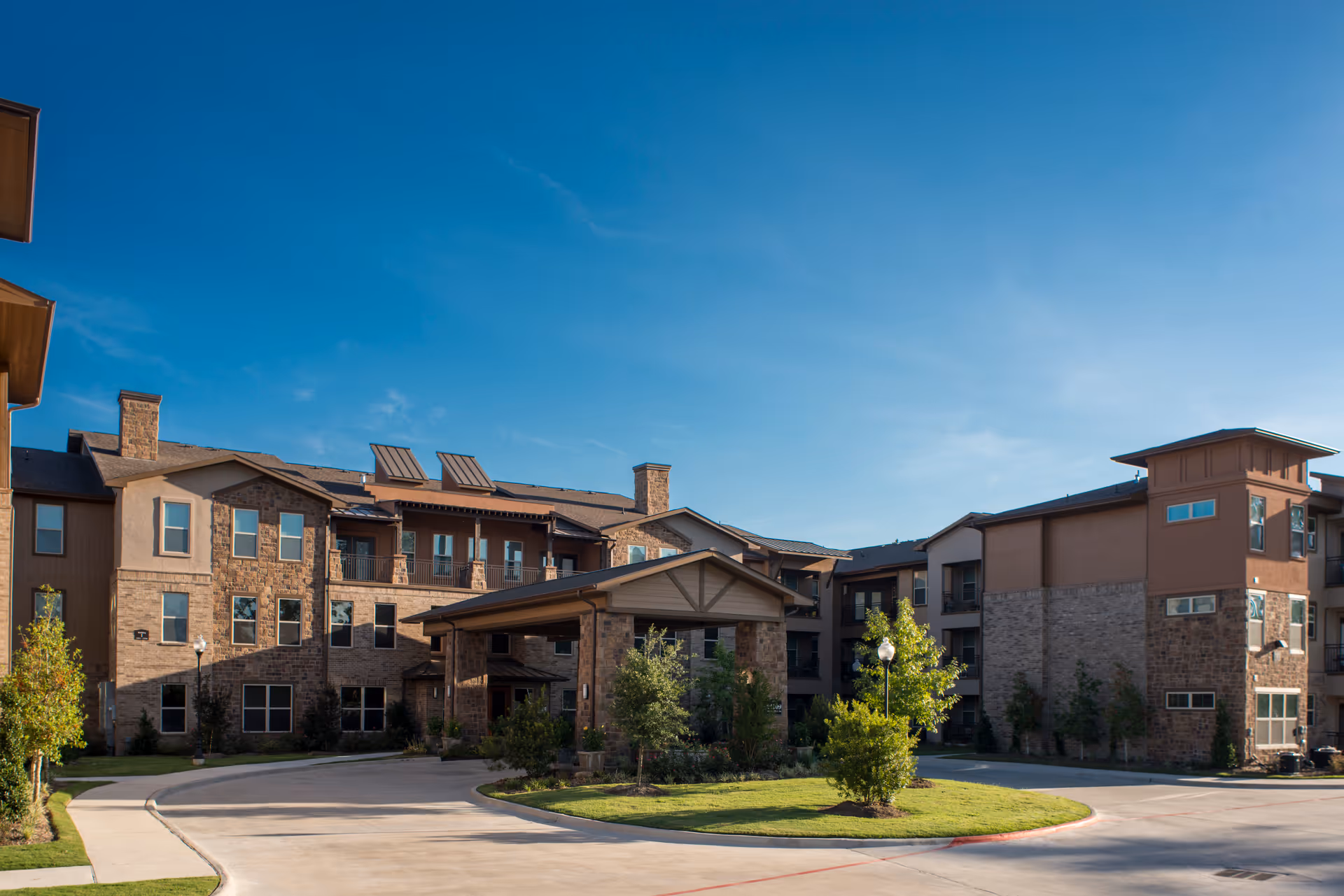 Exterior view of a multi-story senior living facility building with stone and beige siding, a covered entrance, landscaped greenery, and a clear blue sky.
