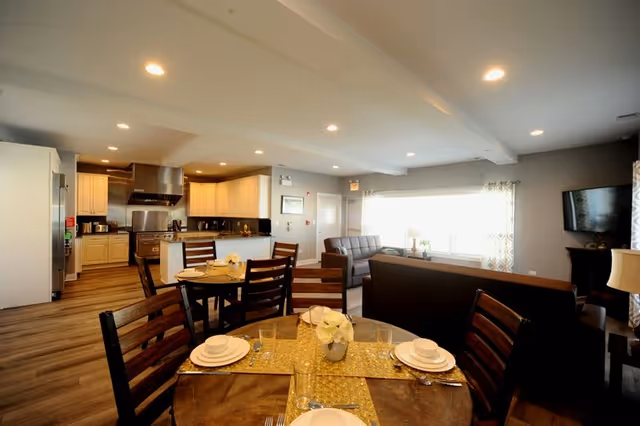 A spacious, well-lit interior featuring a combined kitchen, dining, and living area. The kitchen has white cabinets, stainless steel appliances, and a large stove with a hood. In the foreground, a round dining table is set with plates, glasses, and a centerpiece of white flowers. The living area includes sofas and a wall-mounted TV, with large windows allowing natural light to fill the room.
