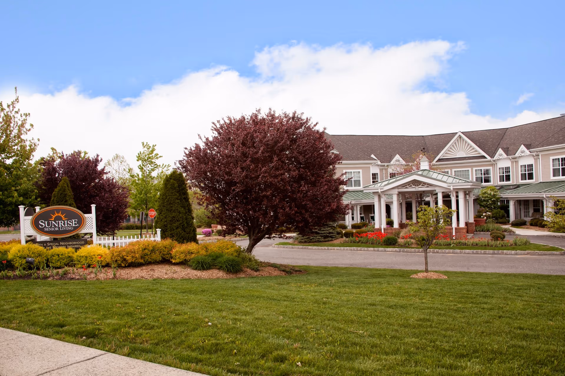 Front entrance of a Sunrise Senior Living building with landscaped lawn, trees, and a facility sign.