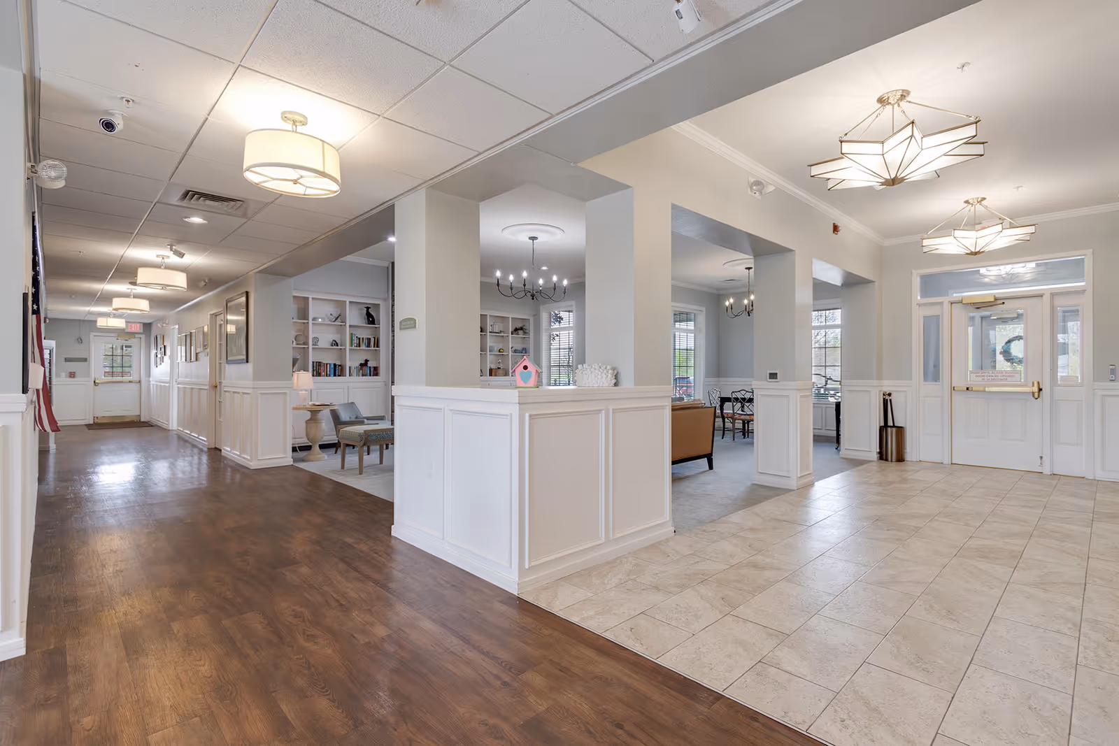 Interior view of a senior living facility hallway with wood flooring on the left and tiled flooring on the right. The hallway leads to a seating area with chairs, bookshelves, and chandeliers. Large windows allow natural light to brighten the space. The area appears clean and welcoming with white walls and ceiling lights.