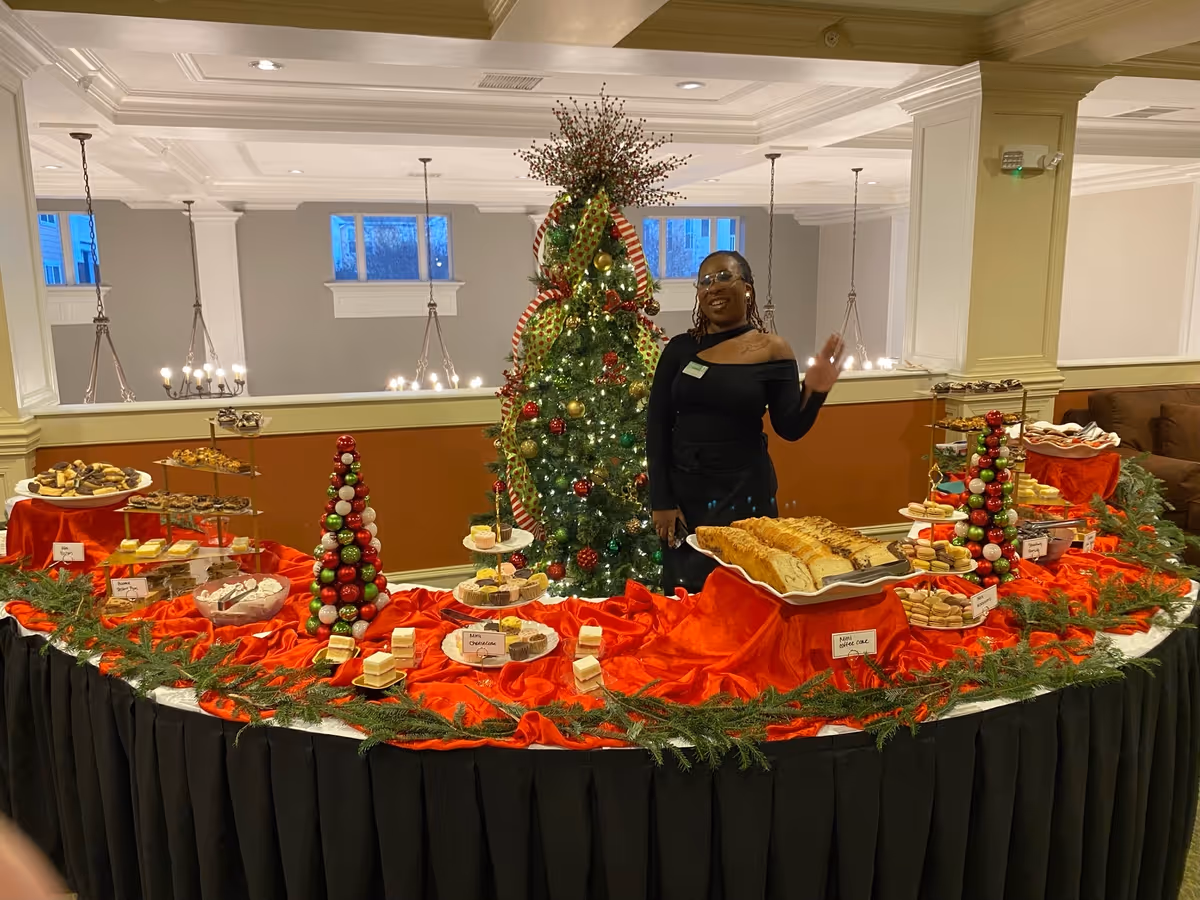 A woman standing behind a festive holiday dessert buffet table decorated with red cloth and green garlands. The table features various desserts including mini cheesecakes, cookies, and pastries. Behind her is a decorated Christmas tree with ornaments and ribbons. The setting appears to be an indoor common area with chandeliers and windows in the background.
