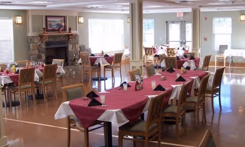 A spacious dining room with multiple tables covered in burgundy tablecloths and white underlays, each set with glasses, napkins, and condiments. The room features wooden chairs, large windows letting in natural light, and a stone fireplace with a framed picture above it.