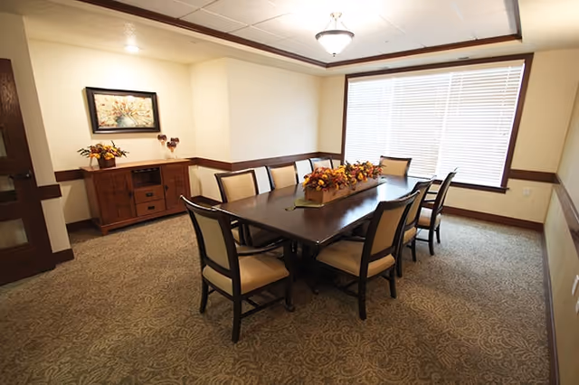 A well-lit dining room featuring a long dark wooden table with upholstered chairs, a floral centerpiece, a sideboard, and a large window with blinds.