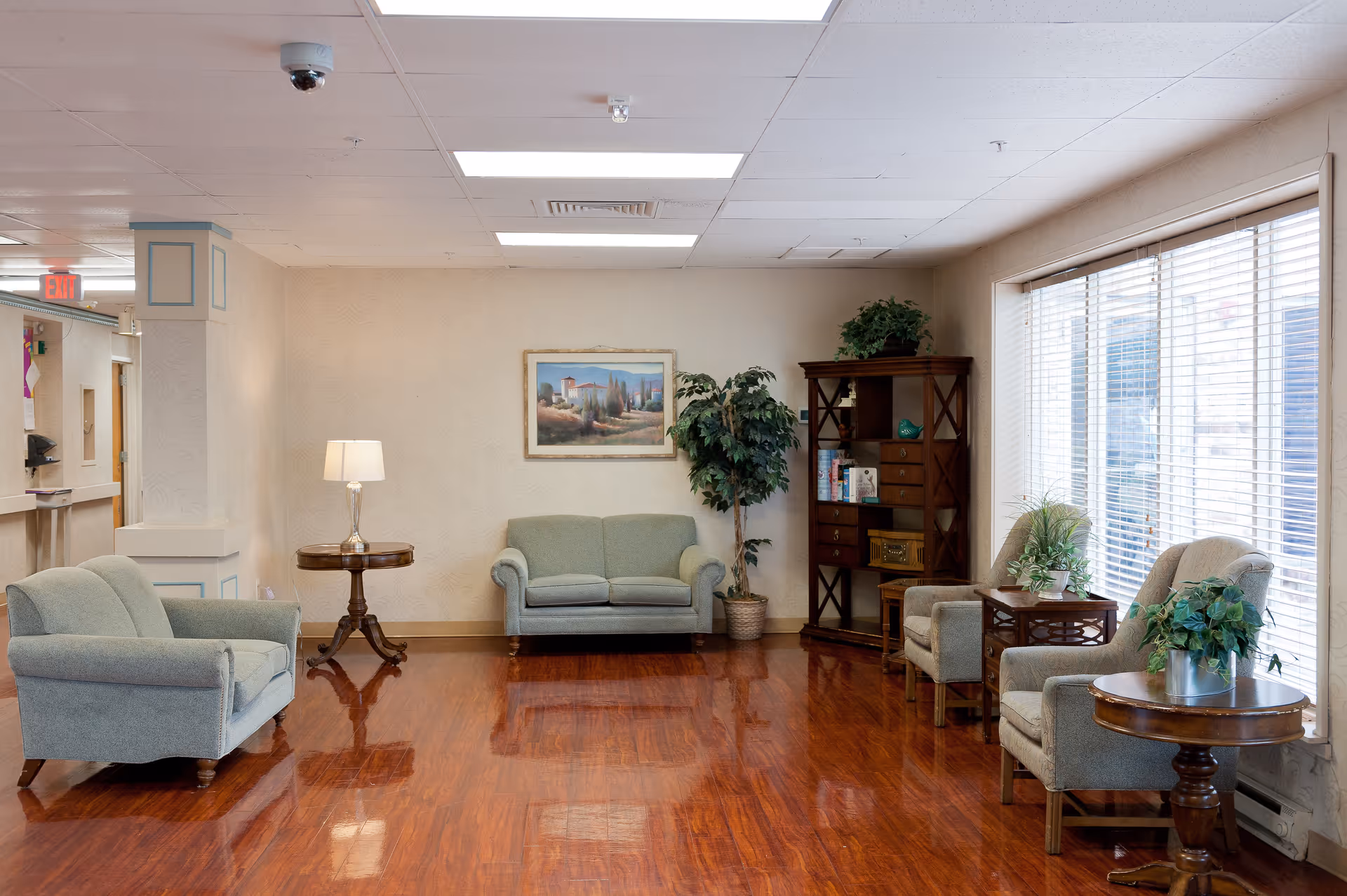A well-lit living room area in a senior living facility with wooden flooring, two light green upholstered loveseats, two matching armchairs, wooden side tables with lamps and plants, a tall wooden bookshelf with decorative items, a large window with blinds, and a framed landscape painting on the wall.