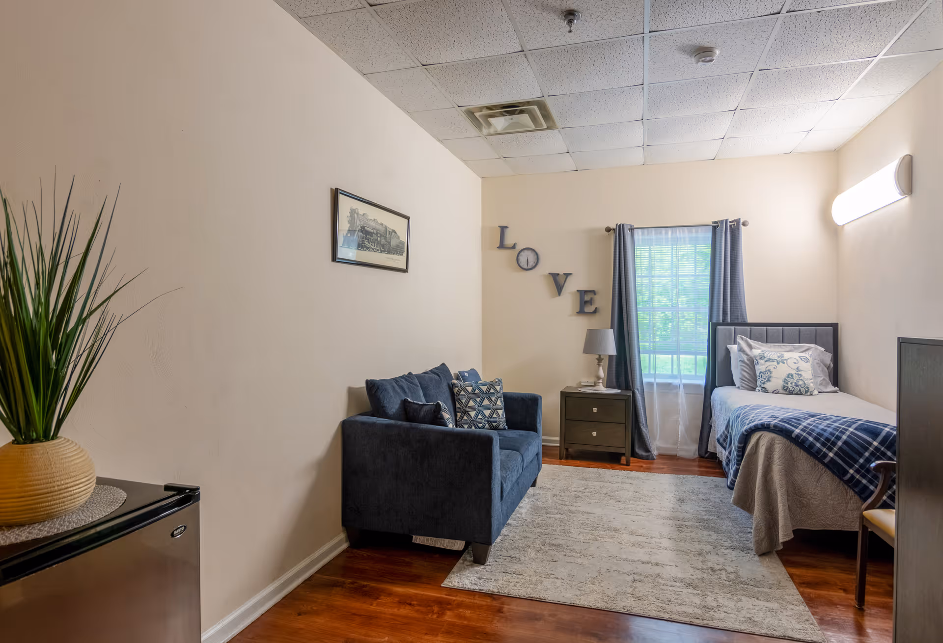 A cozy bedroom in a senior living facility featuring a single bed with pillows and a blue plaid blanket, a dark blue loveseat with patterned cushions, a small nightstand with a lamp, a window with blue curtains, a framed picture of a train on the wall, and decorative letters spelling 'LOVE' with a clock in place of the 'O'. The room has wooden flooring, a light-colored rug, and a ceiling with white tiles.