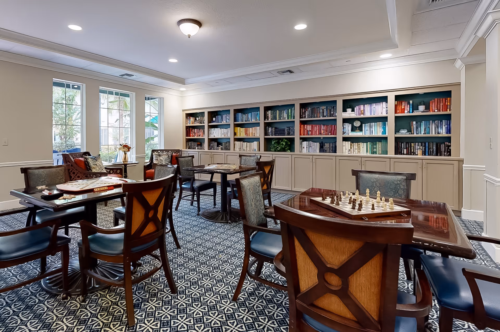 A well-lit common room with multiple wooden tables and chairs arranged for playing board games and chess. The room features a patterned carpet, large windows letting in natural light, and a built-in bookshelf filled with books organized by color along one wall.