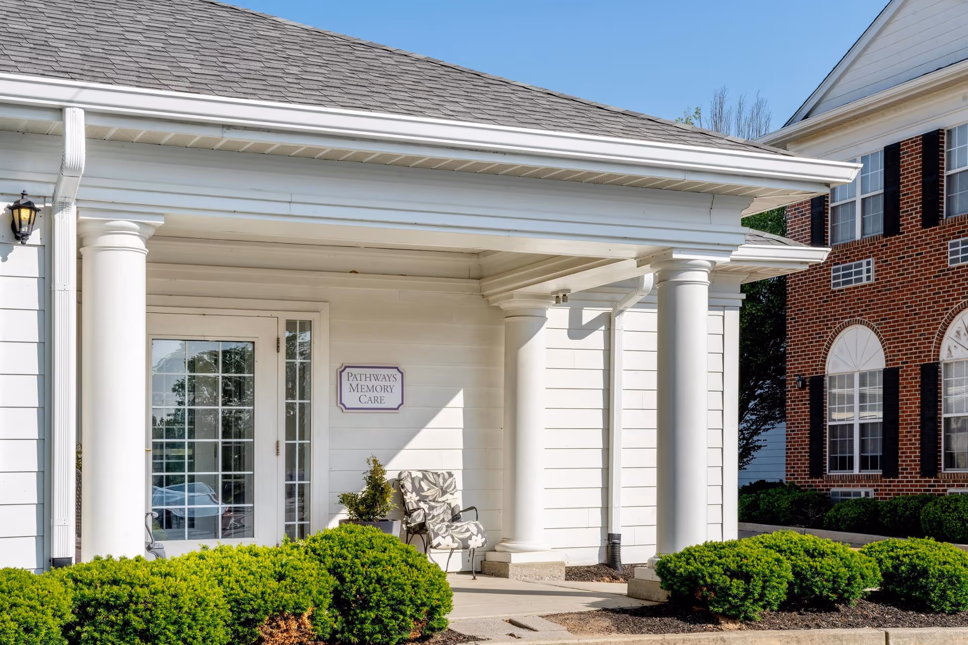 Front entrance of a white-columned memory care building with a glass door, bench, and trimmed shrubbery.