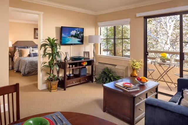 A cozy living room area with a dark wooden coffee table, a blue sofa, and a TV mounted on the wall above a wooden cabinet. There is a large window and a glass door leading to a balcony with a small table and chairs. A bedroom with a neatly made bed is visible through an open doorway. The room is decorated with plants and has beige walls and carpet.