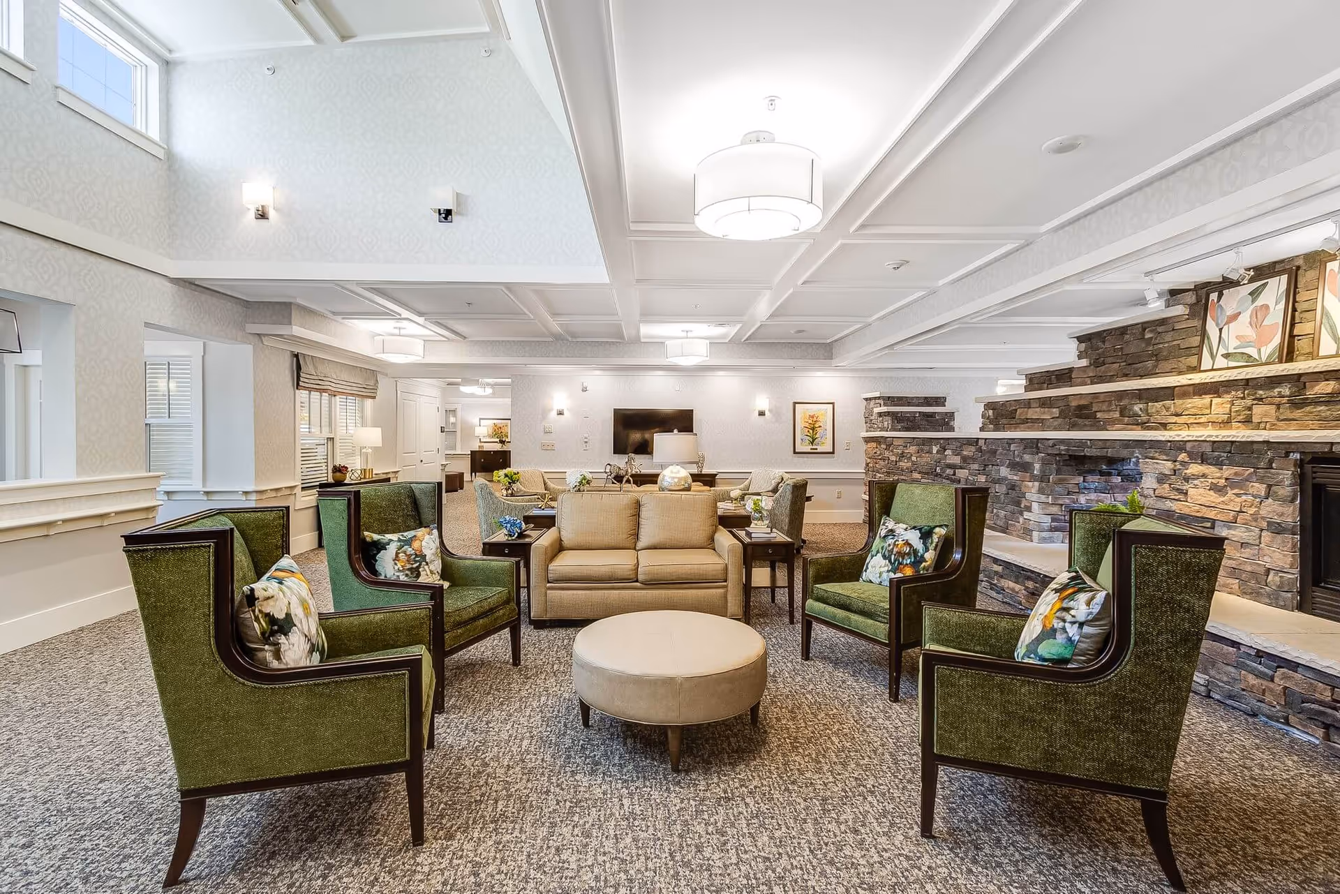 A spacious and well-lit living room area in a senior living facility with a beige sofa, four green armchairs with floral cushions arranged around a round beige ottoman. The room features a stone fireplace on the right side with framed floral artwork above it, patterned wallpaper, carpeted floor, and ceiling lights. There is a TV mounted on the far wall and additional seating in the background.