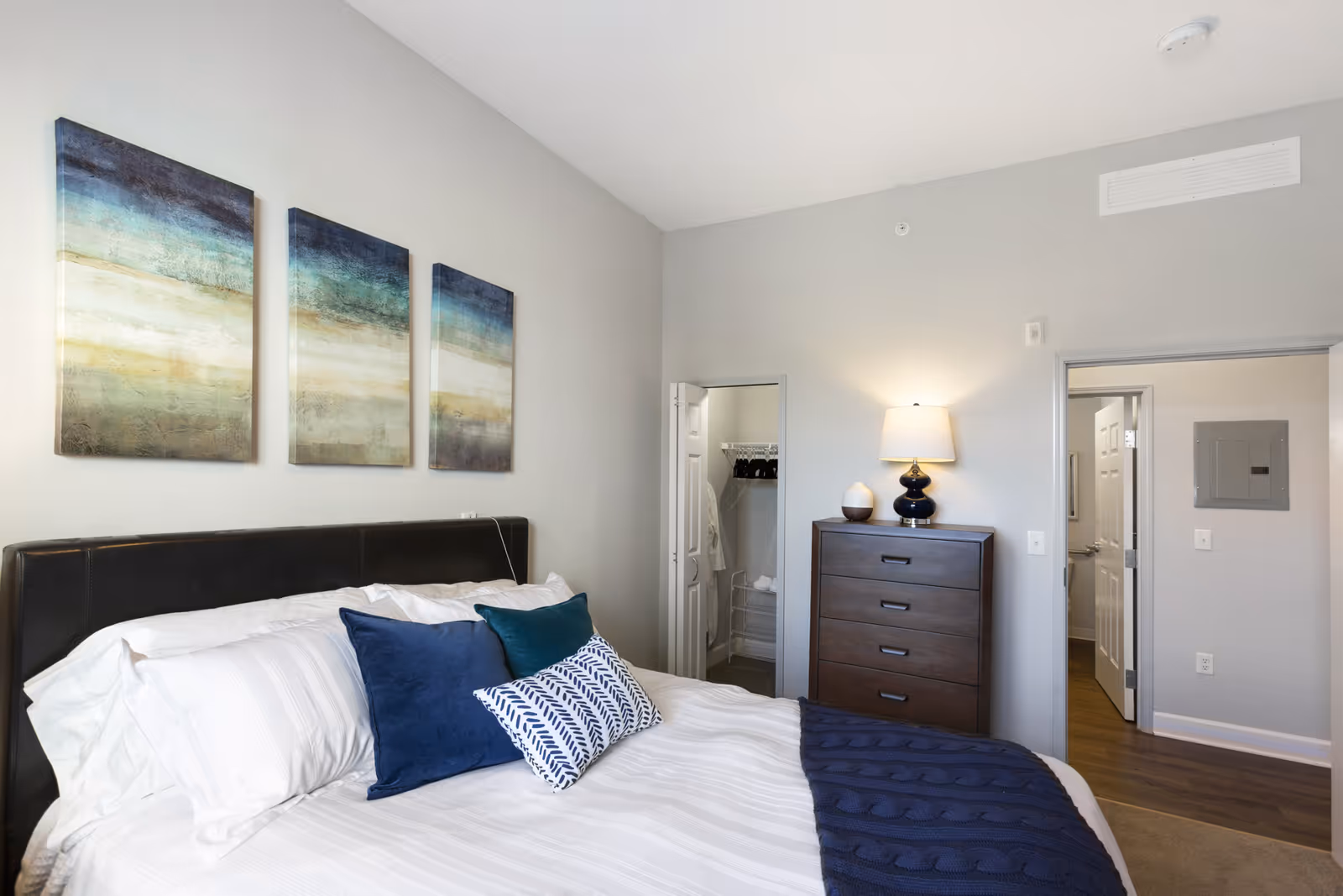 A neatly made bed with white linens and blue accent pillows in a bedroom. Above the bed are three abstract landscape paintings in shades of blue, green, and beige. To the right, there is a dark wooden dresser with a lamp and a decorative item on top. Two doorways are visible, one leading to a closet with hangers and shelves, and the other leading to a hallway with wood flooring.
