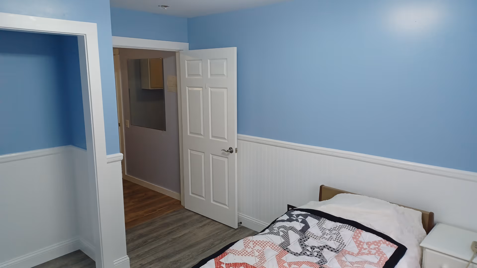 A small bedroom with light blue walls and white wainscoting. There is a single bed with a patterned quilt and a white pillow. Next to the bed is a white nightstand. The door to the room is open, showing a hallway with wood flooring.