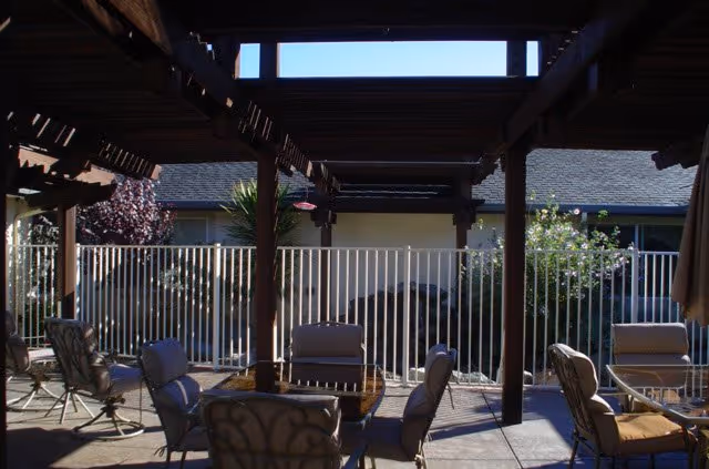 Outdoor patio area with several cushioned chairs and tables under a wooden pergola. There is a white metal fence in the background with shrubs and a building visible behind it.
