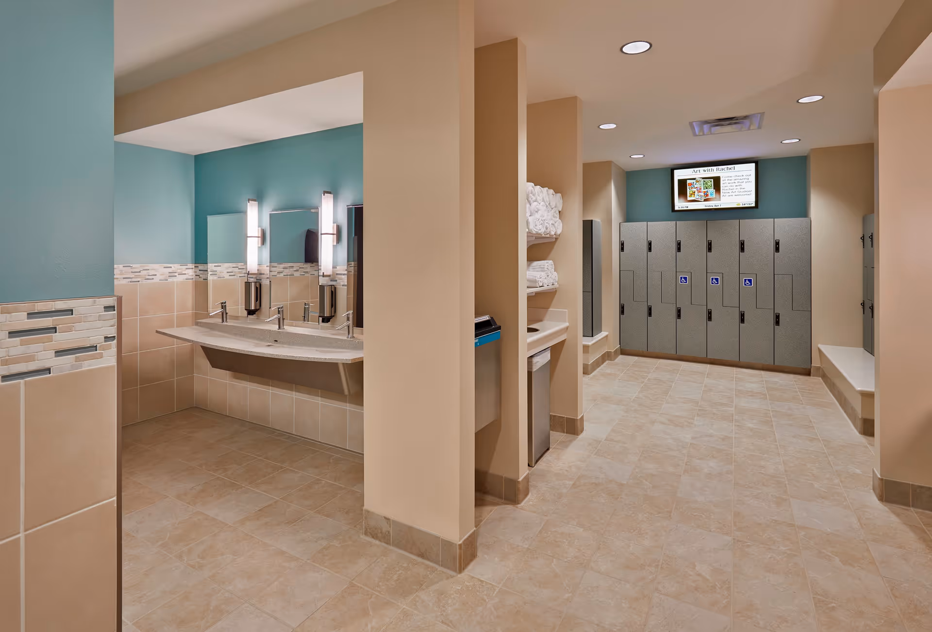 A clean and modern locker room area with beige tiled floors and walls. On the left side, there is a sink area with two faucets, mirrors, and wall-mounted soap dispensers. In the center, there are shelves stocked with rolled white towels and a trash bin below. On the right side, there are multiple gray lockers with handicap accessibility signs and a bench along the wall. A digital screen is mounted above the lockers displaying information.