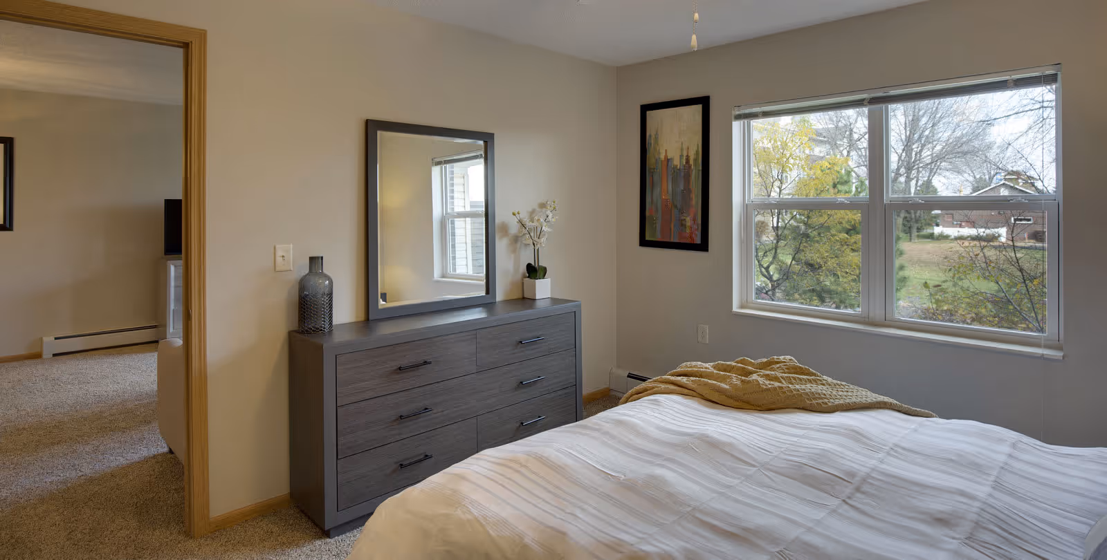 A bedroom with a large window showing an outdoor view with trees and houses. The room has a bed with a white comforter and a beige throw blanket. There is a gray dresser with six drawers against the wall, topped with a decorative vase and a small potted plant. Above the dresser is a rectangular mirror. A framed abstract painting hangs on the wall next to the window. The bedroom opens into a carpeted living area with a TV and a couch visible.