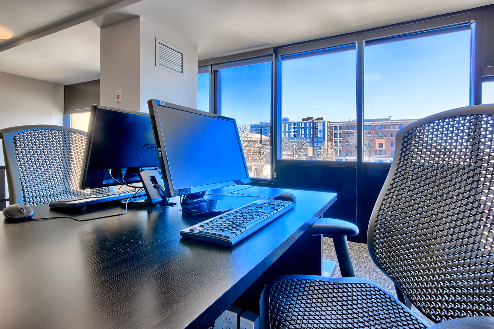 Modern office workspace with two computer monitors, a keyboard, and a mouse on a black desk. Two mesh office chairs are positioned at the desk. Large windows in the background provide a view of buildings and a clear blue sky outside.