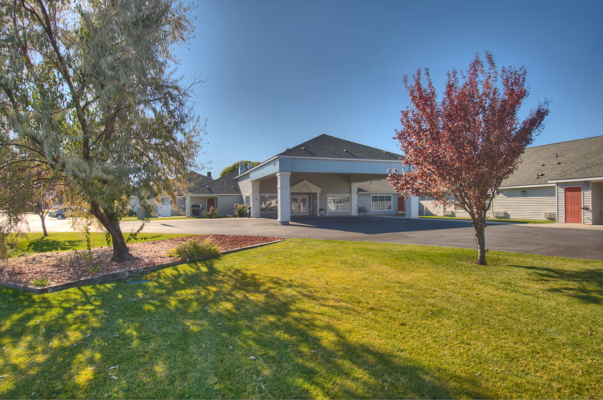 Exterior view of Dorian Place Assisted Living facility showing a single-story building with a covered entrance, surrounded by a well-maintained lawn, trees, and a clear blue sky.