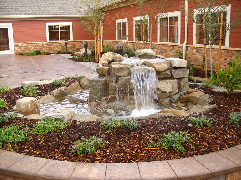 A landscaped outdoor area featuring a small rock waterfall fountain surrounded by mulch and green plants, with a building wall and windows in the background.