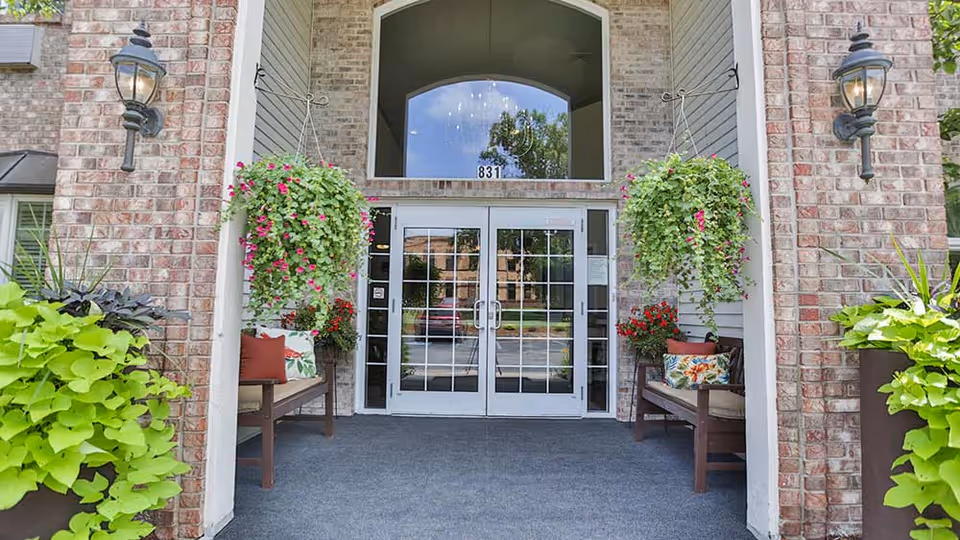 Entrance to a senior living facility with double glass doors framed by brick walls. Two hanging flower baskets with green leaves and pink flowers flank the doorway. There are benches with colorful cushions on either side of the entrance, and two wall-mounted lantern-style lights are visible on the brick walls.