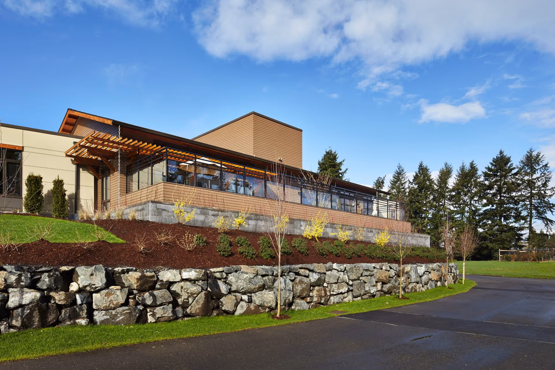 Exterior view of a modern building with large windows and a wooden pergola, surrounded by landscaped greenery, stone retaining walls, and a paved driveway under a partly cloudy blue sky.