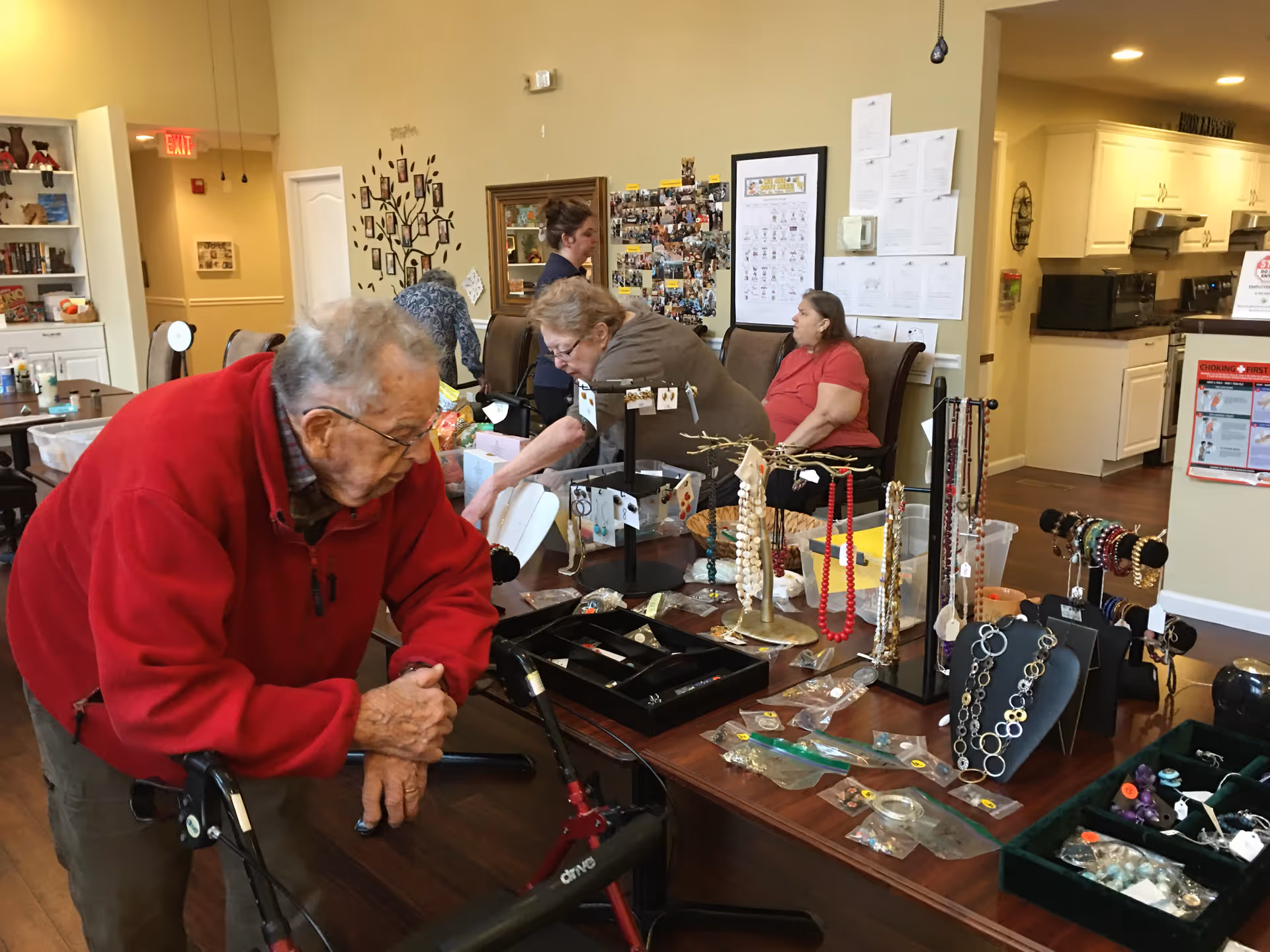 An elderly man in a red jacket leans on a walker while looking at a table displaying various jewelry items including necklaces, bracelets, and earrings in a senior living facility common area. In the background, two elderly women and a staff member are engaged in activities near a wall decorated with photos and artwork. The room has a cozy atmosphere with a kitchen visible in the background.