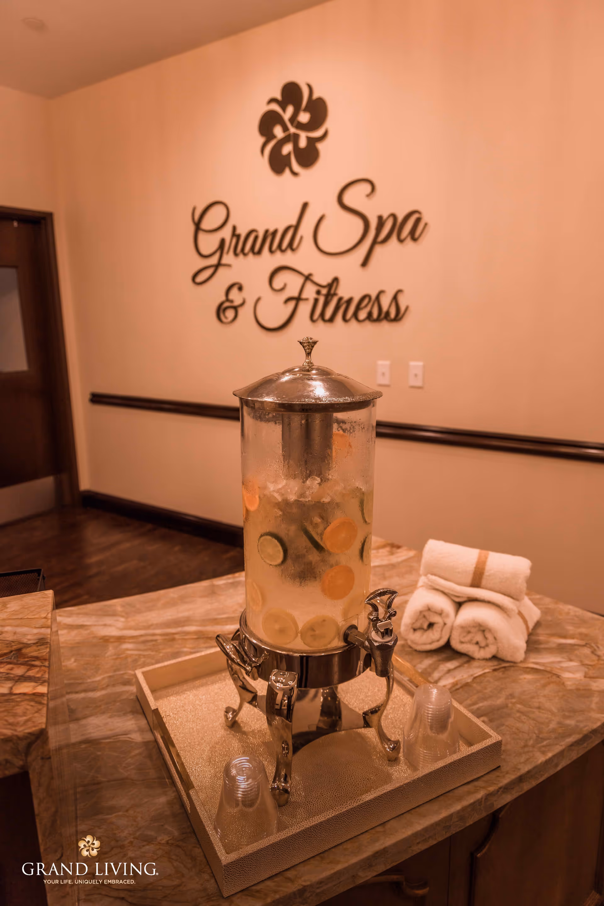 A beverage dispenser filled with water and slices of citrus fruits sits on a marble countertop next to rolled white towels. Behind it, a wall displays the words 'Grand Spa & Fitness' with a decorative flower logo above the text.