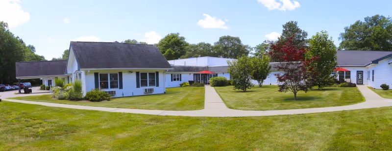 Exterior view of South Kingstown Nursing & Rehab facility showing white buildings with black roofs surrounded by green lawns, trees, and paved walkways under a partly cloudy blue sky.