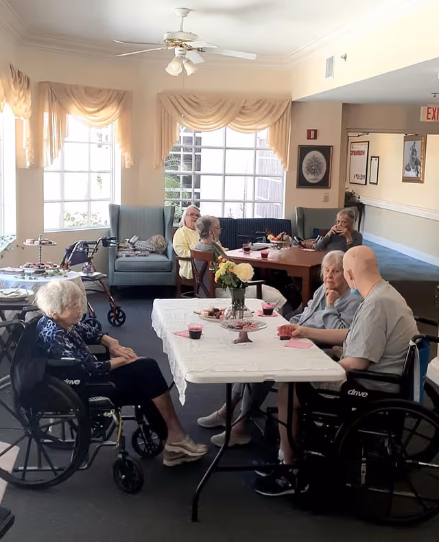 A group of elderly individuals sitting around tables in a well-lit common area with large windows and beige curtains. Some are seated in wheelchairs while others sit in regular chairs. The tables have white tablecloths, flowers, and refreshments. The room has a ceiling fan and framed pictures on the walls.