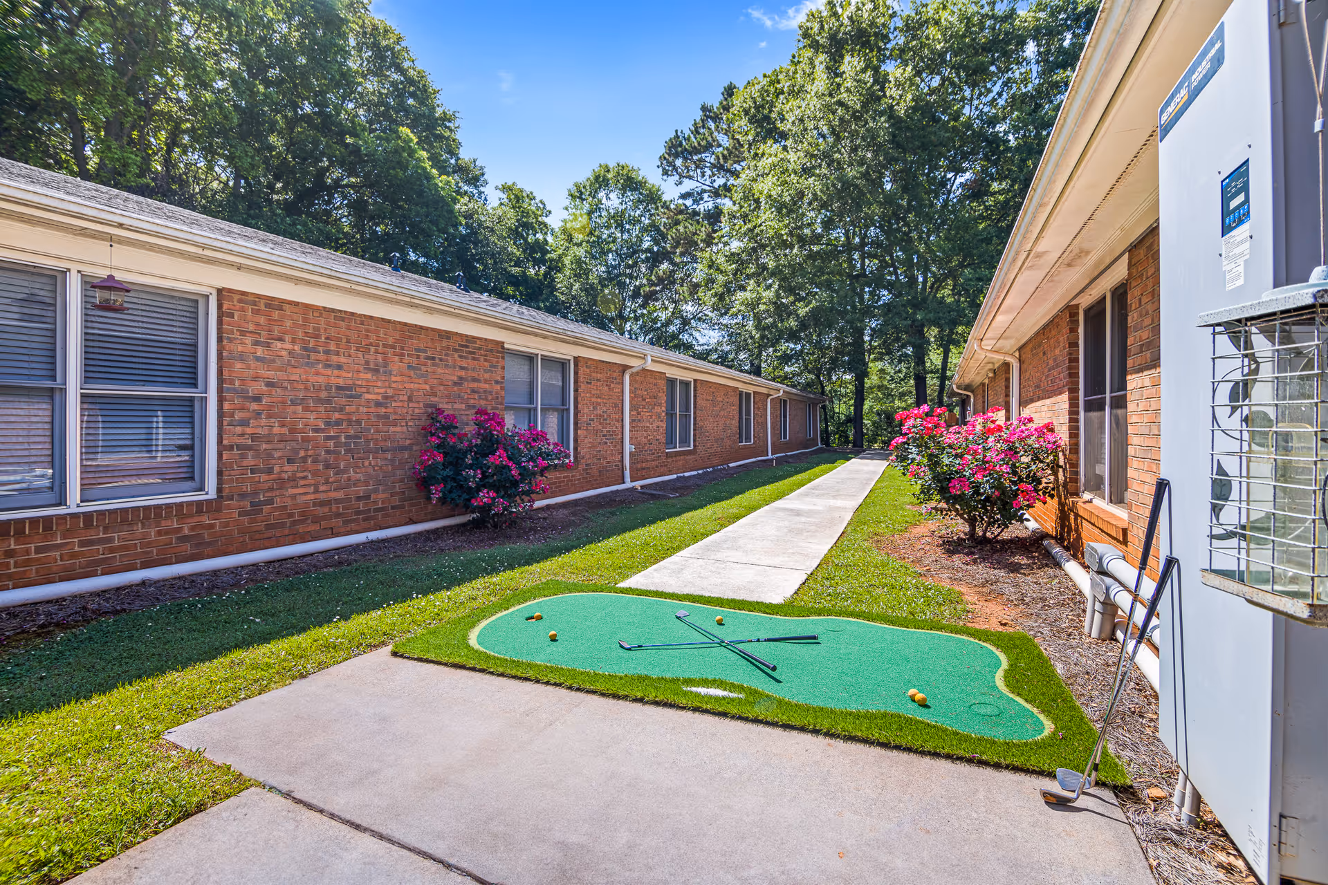 Outdoor view of a pathway between two single-story brick buildings with windows. There is a small artificial putting green with golf clubs and balls on the grass near the sidewalk. Flowering bushes with pink flowers are planted along the buildings, and tall trees are visible in the background under a clear blue sky.