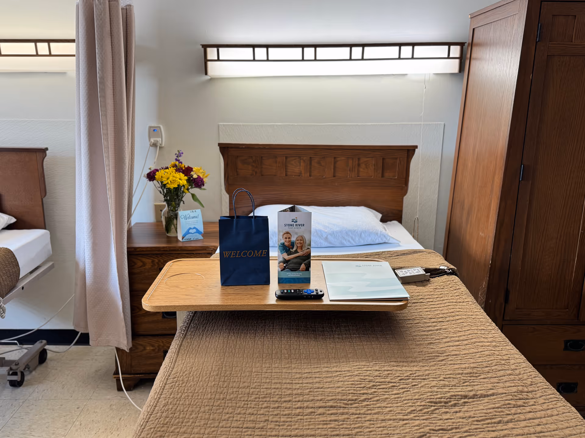A neatly made hospital-style bed with a brown quilted blanket and a wooden overbed table holding a welcome bag, a brochure with a photo of two smiling people, a remote control, and a folder. Behind the bed is a wooden headboard, a wall-mounted light fixture, and a wooden wardrobe. To the left, there is a bedside table with a vase of yellow and purple flowers and a welcome card, partially obscured by a privacy curtain.