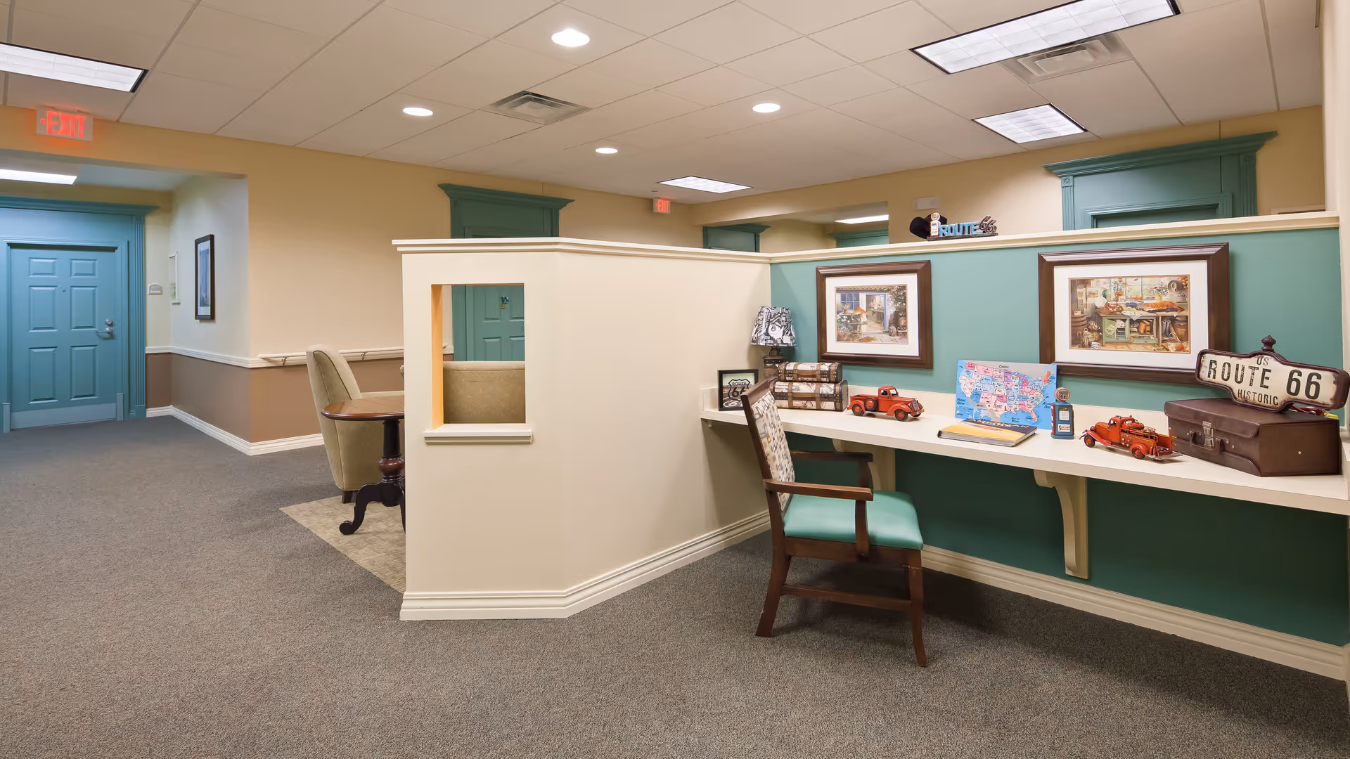 Interior view of a senior living facility hallway with beige and green walls, a carpeted floor, and a built-in desk area decorated with Route 66 memorabilia, framed pictures, and small model cars. There is a wooden chair with a green cushion at the desk, and a seating area with an armchair is visible in the background near a teal door.