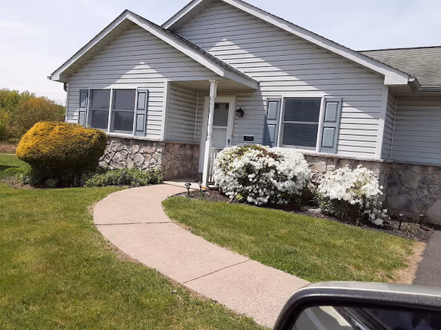 Exterior view of a single-story residential building with light gray siding and stone accents. There is a curved concrete walkway leading to a white front door, flanked by two windows with blue shutters. The landscaping includes green grass, a trimmed bush on the left, and white flowering shrubs near the entrance. The image is taken from a vehicle, with a side mirror visible in the lower right corner.