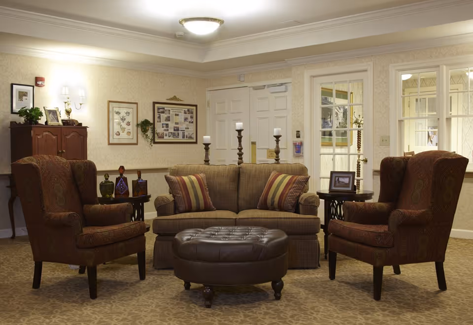 Comfortable lounge area with a sofa, two upholstered armchairs, a leather ottoman and side tables in a senior living facility.