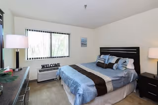 A bright bedroom with a large window, a bed with blue and brown bedding, two bedside tables with lamps, a dresser with a lamp and some decor, and a 'WELCOME' sign on the air conditioning unit below the window.