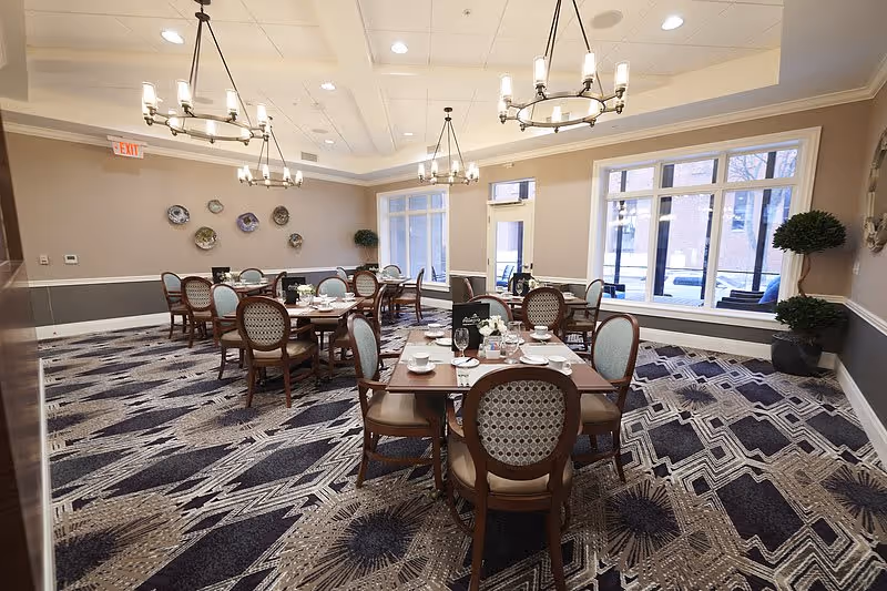 A well-lit dining room with several round tables set with white cups, plates, and silverware. The room features patterned carpet, light-colored walls with decorative plates mounted on one wall, large windows letting in natural light, and elegant chandeliers hanging from the ceiling. There are upholstered wooden chairs around the tables and some potted plants near the windows.