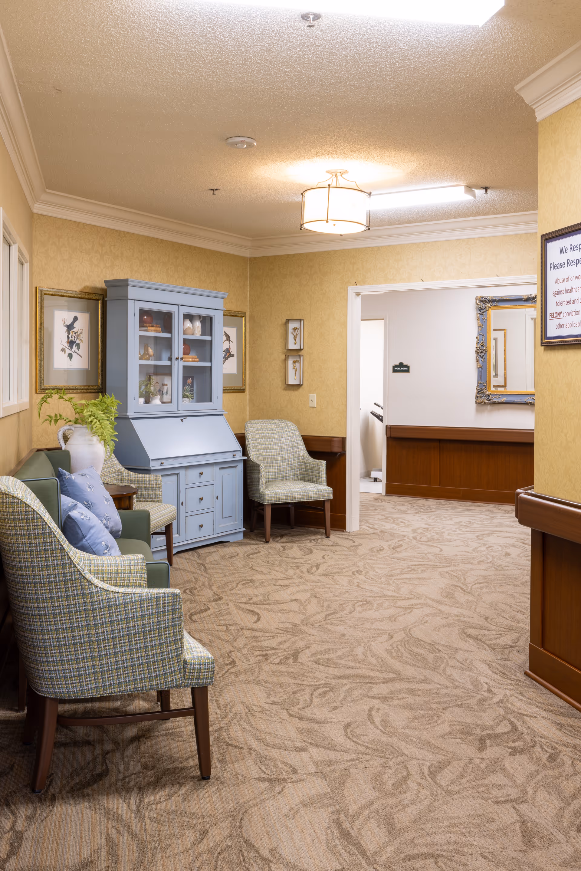 Carpeted seating area in a senior living hallway with upholstered chairs, a light-blue cabinet, framed artwork, and a doorway to another room.