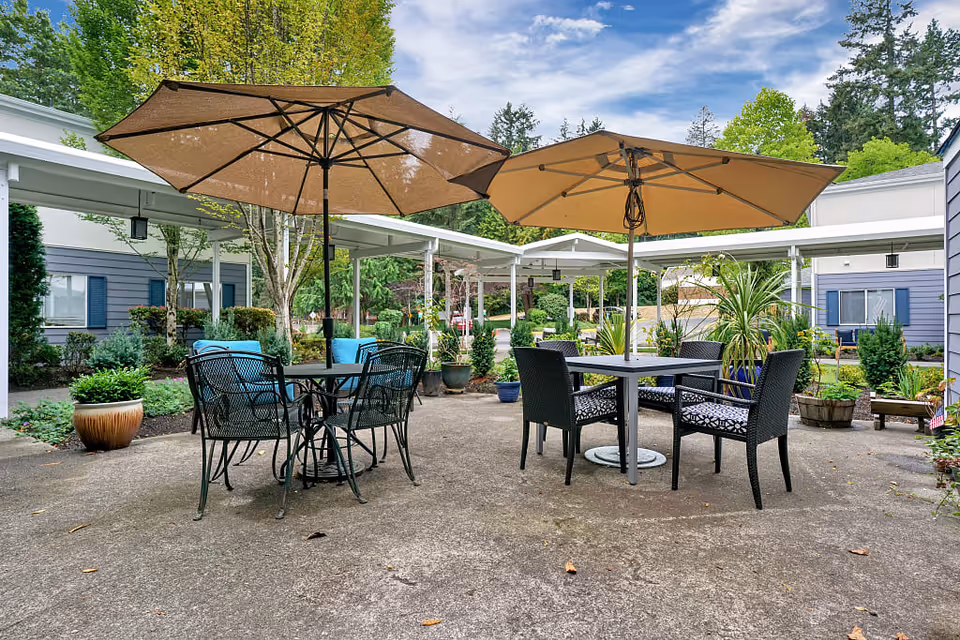 Outdoor patio area with two tables, each shaded by large umbrellas. One table has four black metal chairs with blue cushions, and the other has four black wicker chairs with patterned cushions. The patio is surrounded by greenery, potted plants, and a covered walkway connecting buildings with blue siding and white trim. Trees and a partly cloudy sky are visible in the background.