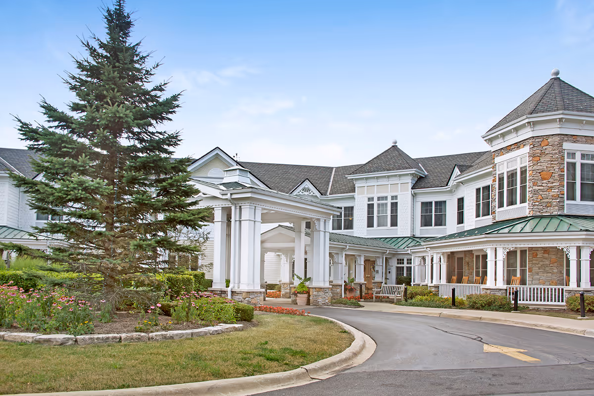 Exterior view of a senior living facility with a large covered entrance, stone and white siding walls, multiple windows, a green metal roof, and a landscaped garden with a tall evergreen tree and flowers in front.