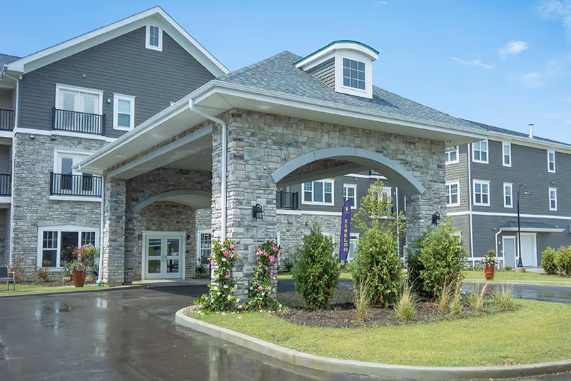 Exterior front view of The Echelon of Medina senior living facility showing a large covered entrance with stone pillars, landscaped greenery, and a clear blue sky.
