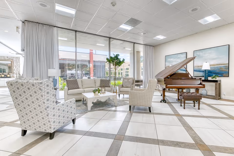 A bright and spacious living room area with large floor-to-ceiling windows. The room features a white sofa, patterned armchairs, a white coffee table, a grand piano with a matching bench, two large landscape paintings on the wall, a console table with a lamp and plant, and a potted plant near the windows. The floor is tiled with a geometric pattern.