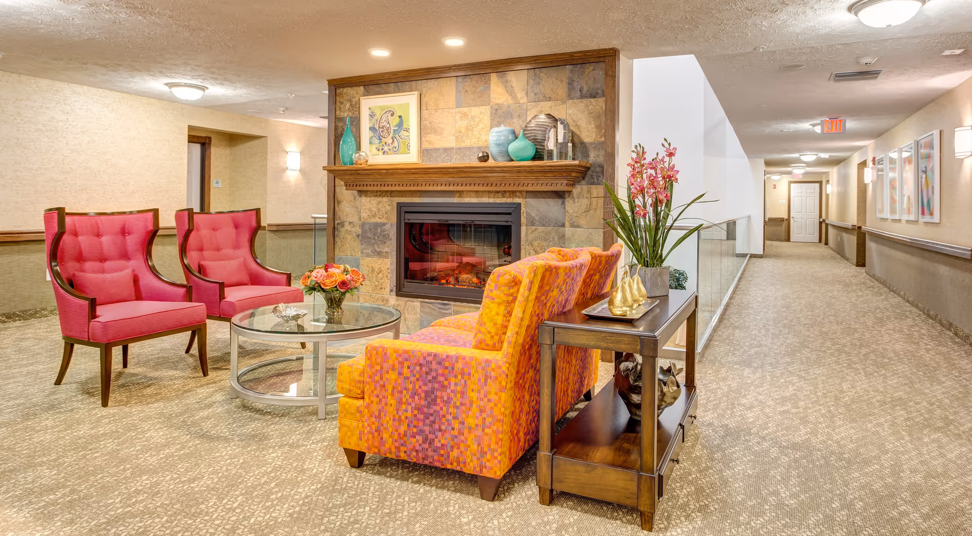 A cozy seating area in a hallway of Quarry Ridge Retirement Community featuring two bright pink armchairs and an orange patterned loveseat arranged around a glass coffee table with a flower vase. Behind the seating area is a decorative fireplace with a wooden mantle adorned with vases and artwork. The hallway extends to the right with beige walls, carpeted floors, and framed artwork on the walls.