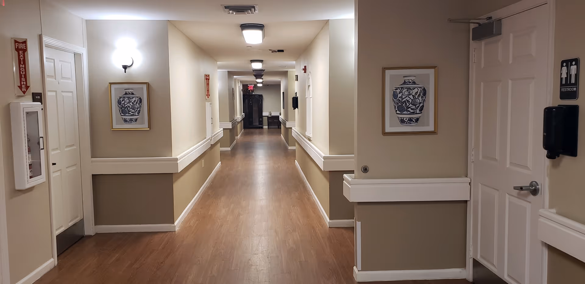 A long, well-lit hallway in an assisted living facility with beige walls and wooden flooring. The hallway has handrails on both sides, framed artwork of blue and white vases on the walls, and several doors including one marked as a restroom. There are ceiling lights evenly spaced along the corridor and a fire extinguisher mounted on the wall.