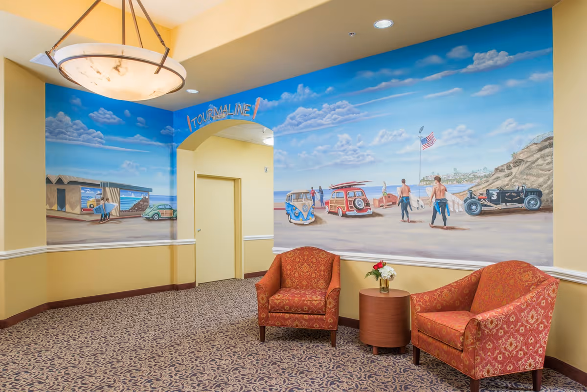 A cozy seating area in a hallway with two red patterned armchairs and a small round wooden table with a flower arrangement. The walls feature a large mural depicting a beach scene with surfers, vintage cars, and an American flag under a blue sky with clouds. A hanging light fixture is visible on the ceiling.